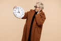 Shocked young hipster woman with blonde short hair wearing a coat and sunglasses holding a clock over beige background. Royalty Free Stock Photo