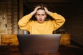 Shocked young male freelancer working online on laptop sitting at a table in a wooden house, looking at the screen and holding Royalty Free Stock Photo