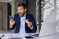 Shocked young man in business suit sitting at desk in office, holding credit card in hand and looking surprised at Royalty Free Stock Photo