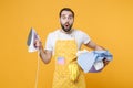 Shocked young man househusband in apron hold basket with clean clothes, iron doing housework isolated on yellow