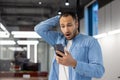 Shocked young muslim man standing in office and holding hand to head, looking at phone screen frustrated and upset Royalty Free Stock Photo