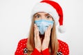 Shocked young woman wearing a Santa Claus hat and a medical protective mask on her face, on a white background, Celebrating New Royalty Free Stock Photo