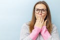 Shocked young woman in eyeglasses covering her mouth with her hands and looking at camera on blue background Royalty Free Stock Photo