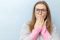 Shocked young woman in eyeglasses covering her mouth with her hands and looking at camera on blue background Royalty Free Stock Photo