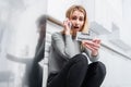 Shocked young woman holding card with lettering home inspection and talking on smartphone near broken refrigerator