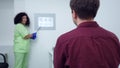 Shooting over shoulder of Caucasian man sitting in ophthalmologic clinic with blurred woman pointing at chart at