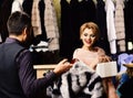 Shop assistant with beard and mustache shows fur coat to lady. Girl in pink fur coat looks at striped fur in male hands