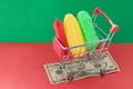 A shopping basket on a red and green background with plastic vegetables inside stands on a banknote. The concept of unnatural