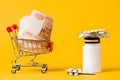A shopping cart filled with cash and a jar of pills. Yellow background. Close up. Concept of high cost of medicines