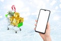 Shopping cart with gift boxes and smartphone mockup, Christmas decorations on background. Christmas online shopping concept Royalty Free Stock Photo