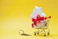 Shopping cart with small white house decorated red bow ribbon and bunch of keys on yellow background. Buying a new home, gift. Royalty Free Stock Photo