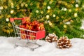 Shopping for Christmas presents. Closeup of red shopping cart and many small gifts in snow with pine cones, fir tree and bokeh Royalty Free Stock Photo