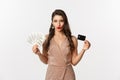 Shopping concept. Sassy woman in elegant dress showing credit card with money, looking pleased at camera, standing over Royalty Free Stock Photo