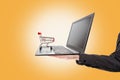 Shopping. A man`s hand holds a laptop with a mini grocery cart standing on it. Orange background. The concept of online shopping Royalty Free Stock Photo