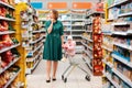 Shopping in the store. A young pensive mother rolls a grocery cart with a baby sitting in it. View among the supermarket shelves.