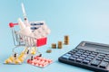 Shopping trolley cart filled pills, tablets and syringe, calculator and coins on blue background, selective focus Royalty Free Stock Photo