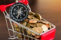 Shopping trolley with gold coins and compass on wood background
