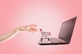 Shopping. A woman`s hand reaches for a mini grocery cart on her laptop. Pink background. Copy space. The concept of online Royalty Free Stock Photo