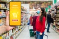 Shopping. A young woman in a medical mask on her face looks at the food displays in the supermarket. The hand holds the smartphone