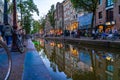 Shops and businesses in red light district reflected in calm canal taken at dusk with long exposure blurring movement by people