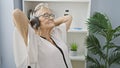 Short-haired, glasses-wearing senior woman boss chilling out in the office, her hands on head, feeling the music through her