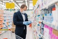 Shot of adult man chooses detergent in housekeeping store, reads label and instruction how to use product, wears medical mask and