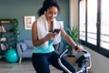 Afro young fitness girl using mobile phone while training on exercise bike at home