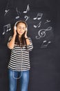 A shot of attractive young woman standing in front of a blackboard with musical notes drawn on and listening to music on