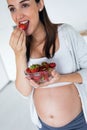 Beautiful pregnant young woman eating strawberries out of bowl in the kitchen at home Royalty Free Stock Photo