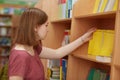 Shot of beautiful young female student stands at college library, chooses book to read, likes study, carries rucksack, dressed in Royalty Free Stock Photo