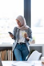 Beautiful young muslim business woman wearing hijab using her smart phone while standing next to the window in the office Royalty Free Stock Photo