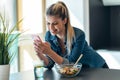 Beautiful young woman eating a bowl of salad while using her mobile phone in the kitchen at home Royalty Free Stock Photo