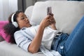 Beautiful young woman listening to music with headphones while using her smartphone sitting on sofa at home Royalty Free Stock Photo