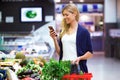 Beautiful young woman looking shopping list on mobile phone while buying fresh vegetables in the market. Royalty Free Stock Photo