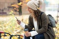 Beautiful young woman sending messages with her smart phone while drinking coffee sitting on a bench in the park in autumn Royalty Free Stock Photo