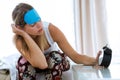 Beautiful young woman sitting on bed with sleep mask trying to wake up with alarm clock in bedroom at home. Royalty Free Stock Photo