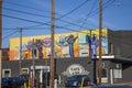 A shot of buildings along a street with a colorful wall mural of hands locked together with power lines and cars and trucks parked