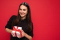 Shot of charming happy smiling brunette girl isolated over red background wall wearing black blouse holding white gift Royalty Free Stock Photo