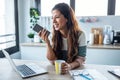 Concentrated young business woman using her mobile phone while working with computer in the kitchen at home Royalty Free Stock Photo