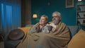 In the shot, an elderly couple is sitting on a sofa under a plaid blanket with a cup of tea or coffee. They are looking Royalty Free Stock Photo