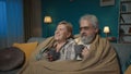 In the shot, an elderly couple is sitting on a sofa under a plaid blanket with a cup of tea or coffee. They are looking Royalty Free Stock Photo