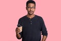 Shot of handsome black man with hesitant expression, has dark skin, wears spectacles, holds takeaway coffee, poses against pink