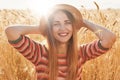 Shot of happy young woman in striped dress and sun hat enjoying sun on wheat field, keeps her hands on head, looking smilling at Royalty Free Stock Photo