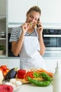 Healthy young woman eating a piece of fresh vegetables and talking with her mobile phone in the kitchen at home.