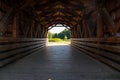 A shot inside a brown covered wooden bridge with a view of lush green trees, blue sky and clouds at the end of the bridge Royalty Free Stock Photo