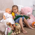 Little girl reads a book with her teddy bear and other toys around her