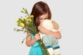 Shot of lovely small female child plays with her favourite toy, holds beautiful flowers, enjoys recieveing present, dressed in