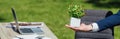 Shot of man holding flowerpot with plant standing in park near table with laptop