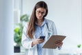 Pretty female doctor using her mobile phone while reviewing medical reports in medical consultation Royalty Free Stock Photo