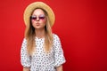 Shot of pretty positive young blonde woman wearing summer dress straw hat and stylish glasses isolated over red Royalty Free Stock Photo
